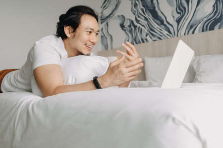 Happy asian man work with laptop on the bed in the hotel. Concept of workation.