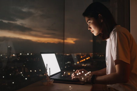 Asian woman work with laptop by the windows apartment with evening city view.の写真素材