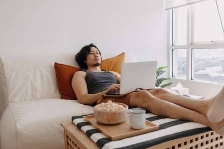 Asian man working at home and eating popcorn in his apartment.の写真素材