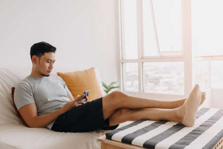 Asian man looking at his mobile while relax on sofa in his living room.の写真素材