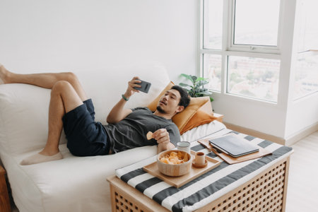 Asian man watch online tv and eating snack instead of working from home.の写真素材