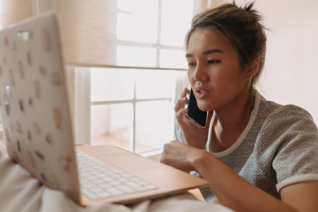 Asian woman talking business on the phone work with her laptop in the bedroom.の写真素材