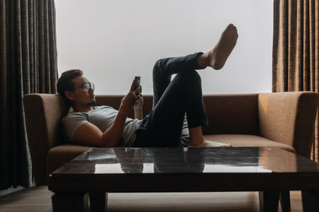 Asian man lies on sofa using phone with his feet on the table. Concept of mannerの写真素材