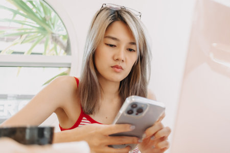 Cute freelance asian girl is using smartphone while working on laptop.