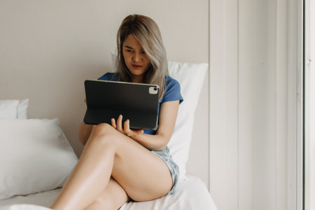 Serious asian woman working online with tablet on hotel bed in travel tripの写真素材