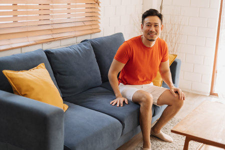 Happy and cheerful asian man in orange t-shirt sit on the sofa in the house.の写真素材
