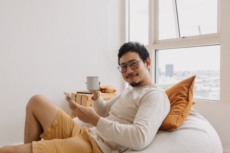 Happy asian man in long sleeve sit relaxed on bean bag using smartphone.