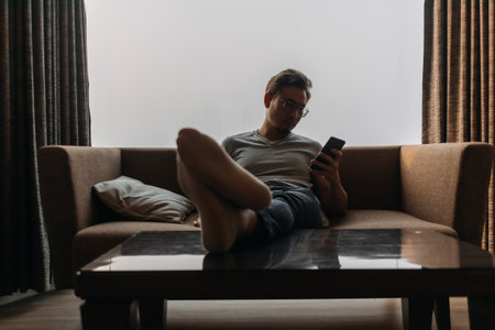 Asian man lies on sofa using phone with his feet on the table. Concept of mannerの写真素材