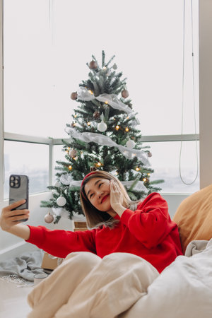 Happy and cheerful Asian woman in red sweater shirt taking selfie photo with her decorated Christmas tree in the apartment in Christmas holiday.の写真素材