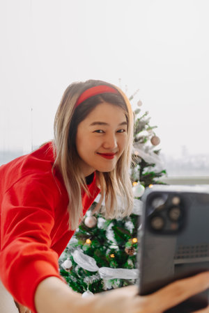 Happy and cheerful Asian woman in red sweater shirt taking selfie photo with her decorated Christmas tree in the apartment in Christmas holiday.の写真素材