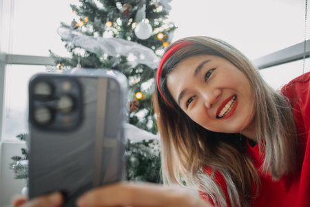 Happy red shirt Asian woman making a video call for holiday greeting with her friends and family showing the Christmas tree. . High quality photoの写真素材