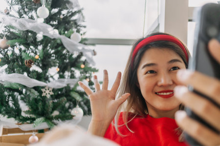 Happy red shirt Asian woman making a video call for holiday greeting with her friends and family showing the Christmas tree. . High quality photoの写真素材