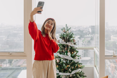 Happy and cheerful Asian woman in red sweater shirt taking selfie photo with her decorated Christmas tree in the apartment in Christmas holiday.の写真素材