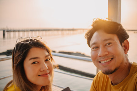 Happy Asian couple lover take selfie photo in the resort with romantic sunset at the sea.の写真素材