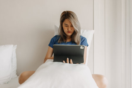 Serious asian woman working online with tablet on hotel bed in travel tripの写真素材