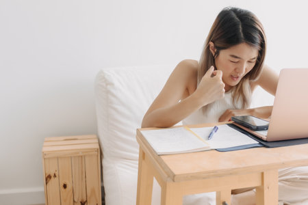 Serious freelance Asian woman is working online at home with computer laptop and taking notes in her...