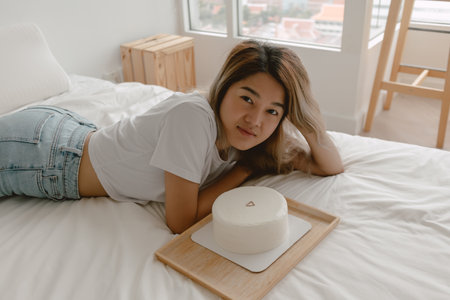 Lovely beautiful happy asian woman lies on the bed admiring her birthday cake.の写真素材