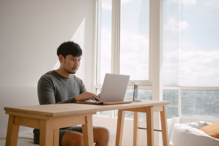 Asian man working with computer in apartment with city view at the windows in morning light.の写真素材