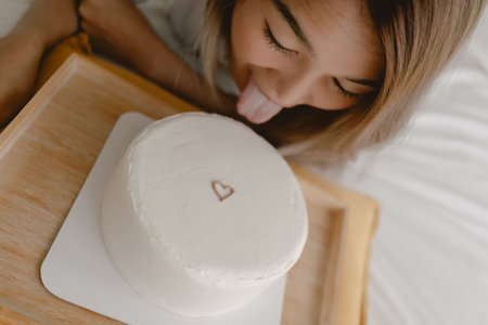 Close up of funny Asian woman tongue out trying to eat white minimal heart cake on the bed.の写真素材