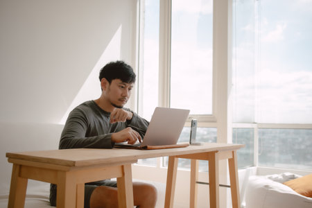 Asian man working with computer in apartment with city view at the windows in morning light.