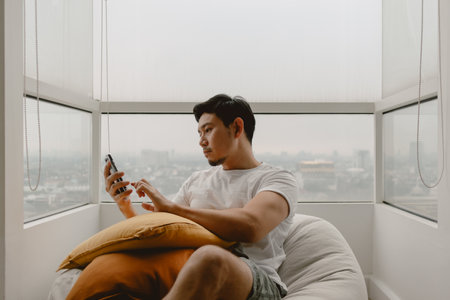 Asian man using mobile phone for killing time with social media online games and chat with friends on the beanbag sofa in apartment.の写真素材