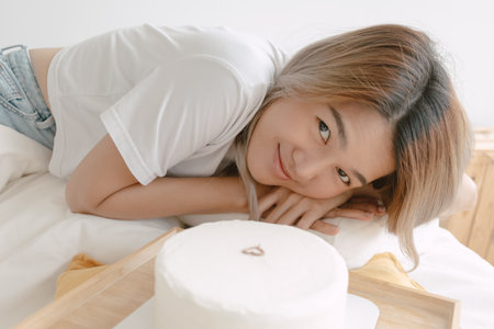 Lovely beautiful happy asian woman lies on the bed admiring her birthday cake.の写真素材