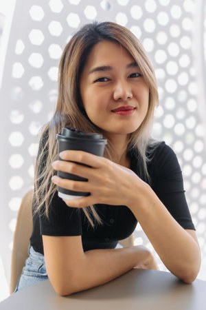 Happy Asian woman in black t-shirt sipping hot coffee in white coffee shop.
