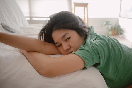 Unhappy asian Thai woman leaning on white cough sofa while resting head on arm with sadness, sitting on wooden floor, thinking something sad, living alone in room apartment.の写真素材