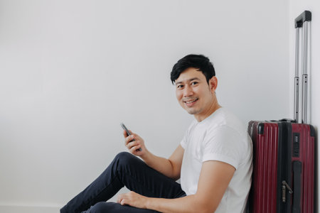 Happy asian man sit on the floor leaning on the suitcase with another hand holding mobile phone.