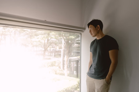 Asian man leaning against white wall in navy blue t-shirt and khaki shorts, looking out of the windows.の写真素材