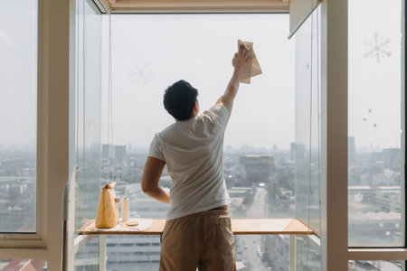 Backside view of asian Thai man wiping window glass in room apartment with city view, keep glass...