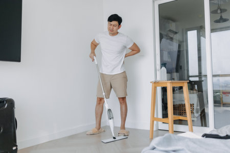 Asian man tired and painful face, standing and holding mop, hand on his waist and back, backache symptom after done housework, mopping the floor. Housework concept.の写真素材