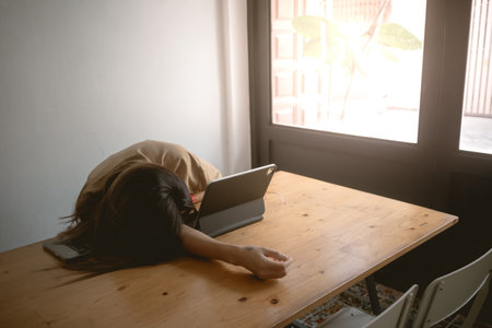 Asian Thai woman taking a nap, fall asleep, resting her head on arm while working and using laptop on wooden table, tired of work sitting on chair.の写真素材