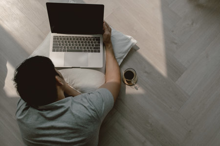 Top view of asian man using computer, lying on floor while working on laptop with coffee and typing...