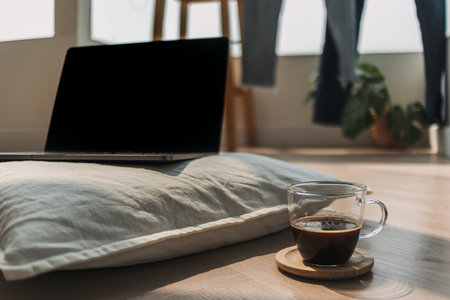 Photo of laptop showing black empty screen, notebook putting on white pillow, on wooden floor in cozy room with black coffee cup.の写真素材