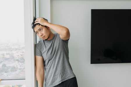 Serious asian man looking at the sky from his apartment, thinking of something.