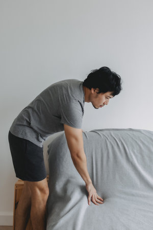 Asian man is doing his house work, covering the sofa with grey sheet.の写真素材