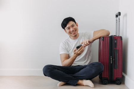 Happy asian man sit on the floor leaning on the suitcase with another hand holding mobile phone.の写真素材
