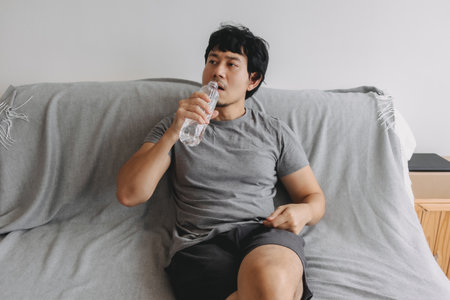 Asian man lifting up the plastic bottle of water to drink, sitting on the sofa.の写真素材