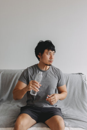 Vertical photo of Asian man drinking water and sitting on the sofa.の写真素材