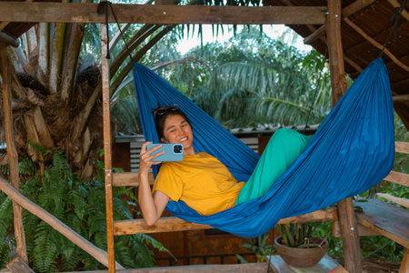Happy asian woman in yellow t-shirt lying on blue hammock taking mobile photo.の写真素材