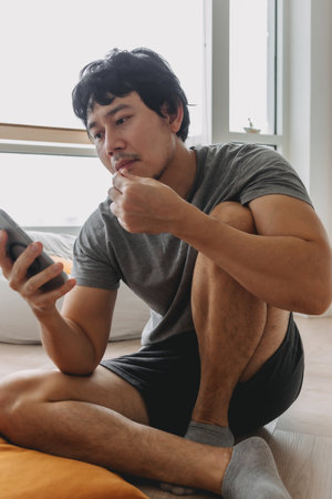 Asian man sitting on the floor eating snacks peanuts and looking at mobile phoneの写真素材