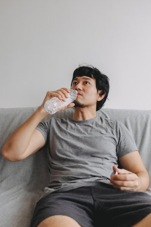 Vertical photo of Asian man drinking water and sitting on the sofa.の写真素材