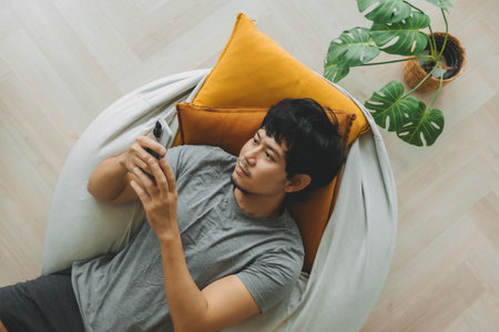 Asian man lean on the bean bag sofa using smartphone in the living room.