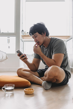 Asian man sitting on the floor eating snacks peanuts and looking at mobile phoneの写真素材
