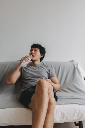 Vertical photo of Asian man drinking water and sitting on the sofa.の写真素材