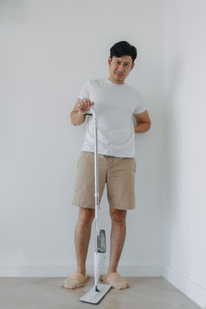 Asian man tired and painful face, standing and holding mop, hand on his waist and back, backache symptom after done housework, mopping the floor. Housework concept.の写真素材