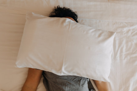 Sleepy asian man keep on sleeping on the bed, using pillow covering his face.の写真素材
