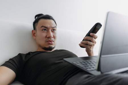 Distracted creative designer Asian man lying on sofa working watching tv.の写真素材