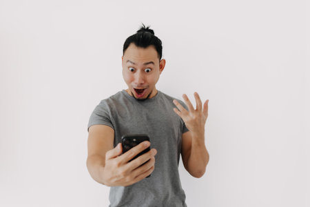 Shock and wow face Asian man in grey t-shirt use smartphone app on white.の写真素材